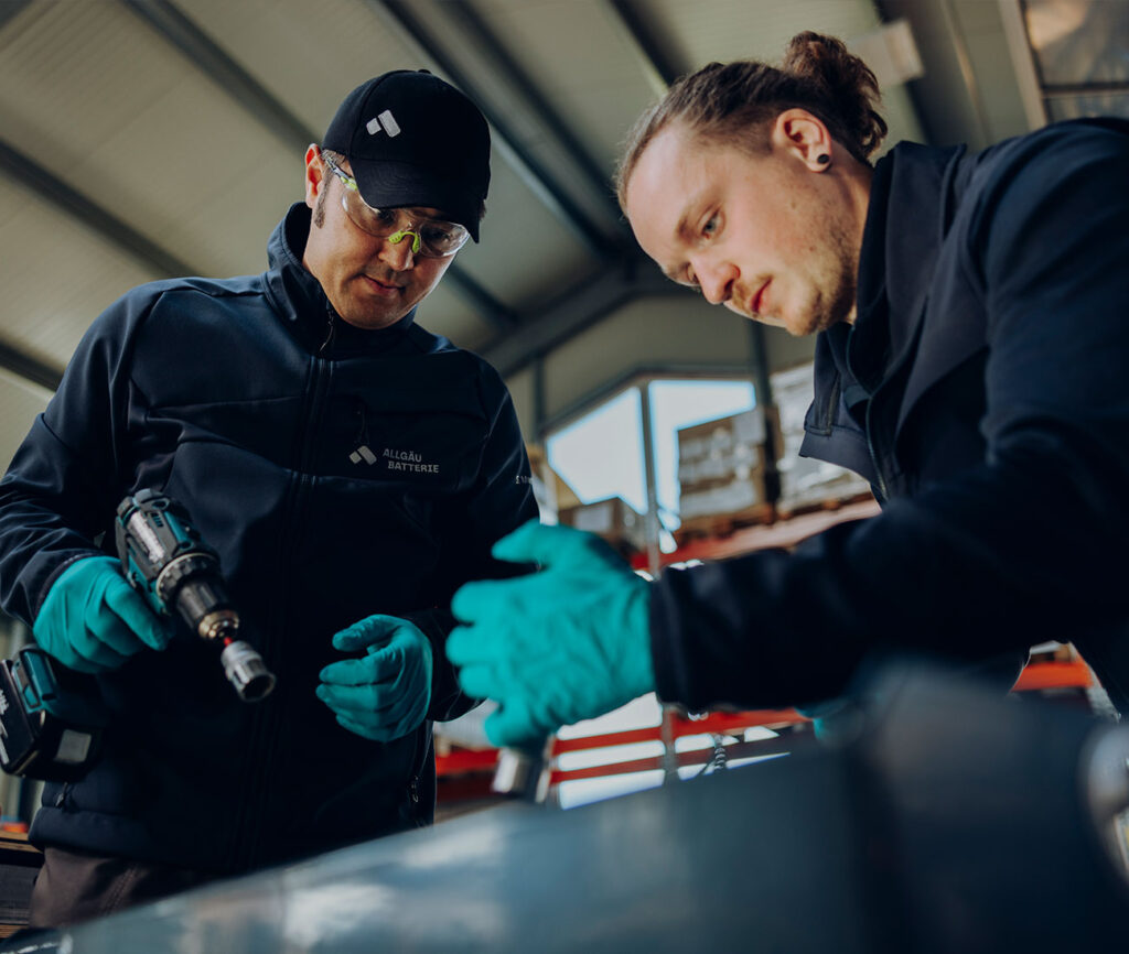 Zwei Mitarbeitende in einer Werkhalle arbeiten konzentriert an einem technischen Bauteil; einer hält einen Akkuschrauber, beide tragen Schutzhandschuhe.