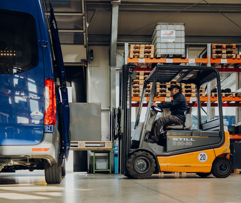 Ein Mitarbeiter fährt einen orangefarbenen Gabelstapler und lädt eine schwere Batterie aus einem geöffneten blauen Servicetransporter in einer Lagerhalle aus.