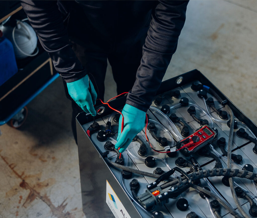 Ein Techniker mit blauen Schutzhandschuhen misst die Spannung einer großen Bleibatterie, auf der ein rotes Multimeter liegt.