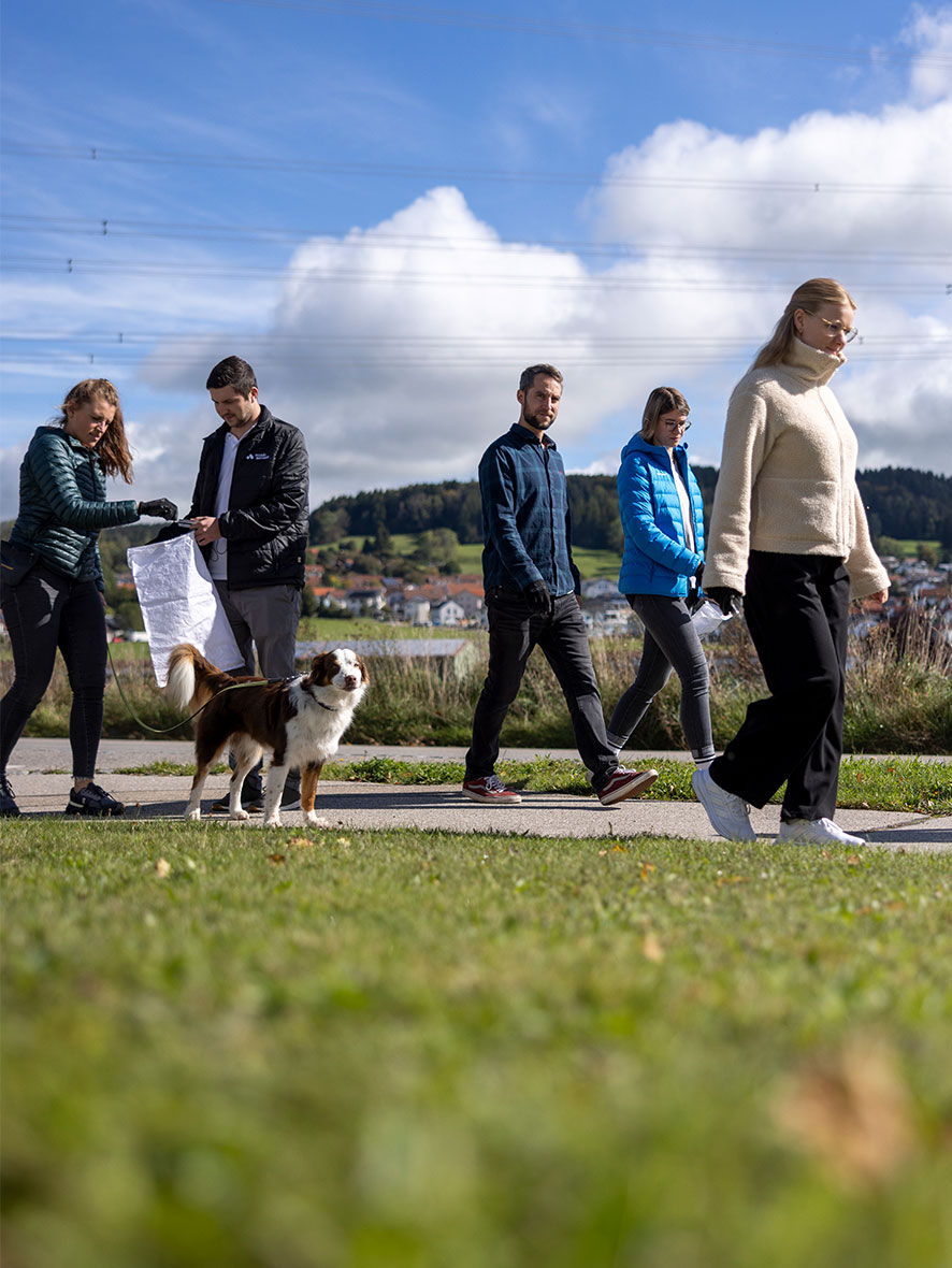 Mehrere Personen gehen bei sonnigem Wetter auf einem Weg durch die Natur und sammeln Müll; ein Hund läuft mit, im Hintergrund sind Wiesen, Hügel und Häuser zu sehen.